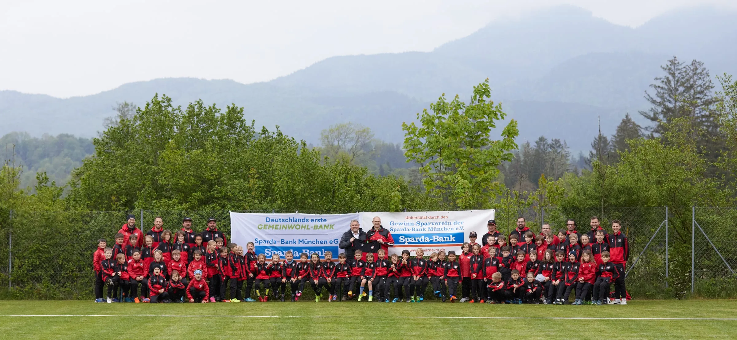 Die Jugendabteilung des SV Hechendorf in roten Vereinsjacken vor einem Sparda-Bank-Banner auf dem Sportgelände, dahinter Bäume und die Murnauer Berge